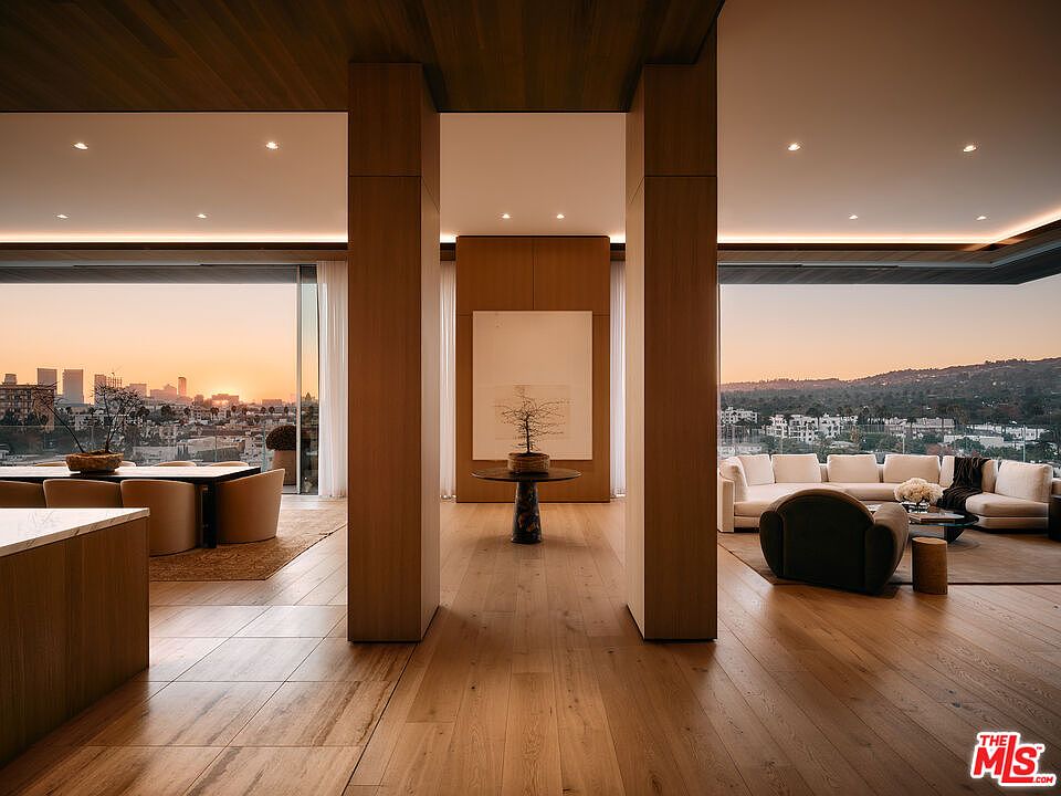 This interior shot showcases a luxurious living space with an open floor plan, featuring hardwood floors and floor-to-ceiling windows offering panoramic city views. The room is divided by architectural columns, with a dining area on one side and a seating area with a white sofa and dark armchair on the other. The overall impression is one of modern elegance and spaciousness.