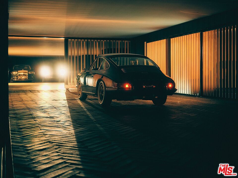 The image showcases a luxurious garage interior featuring a classic black Porsche parked on a herringbone brick floor. In the background, another luxury car with illuminated headlights is visible, adding depth. The garage is illuminated with warm lighting, creating a sophisticated and inviting atmosphere, enhanced by the slatted wall design.