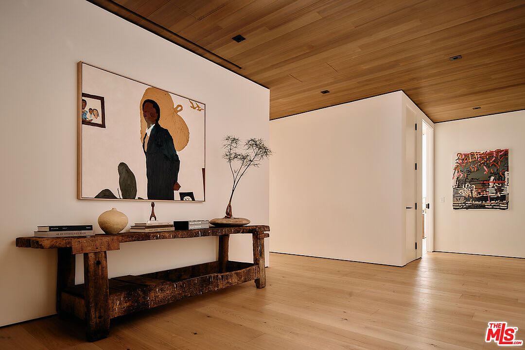 This interior shot showcases a modern hallway with light wood flooring and a wood-paneled ceiling. A rustic wooden console table displays art and decorative objects, while large paintings adorn the walls, adding a touch of sophistication. The space is well-lit and exudes a contemporary, minimalist aesthetic.