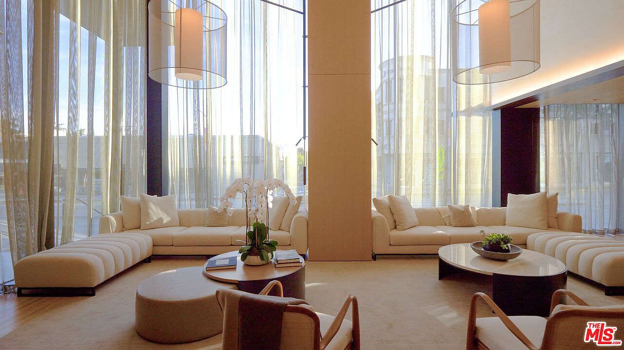 This is an interior shot of a luxurious living room featuring two large, cream-colored sofas with matching ottomans, arranged around round coffee tables. The room is bathed in natural light streaming through floor-to-ceiling windows, which are partially covered by sheer curtains. The overall impression is one of elegance and spaciousness, perfect for a high-end real estate listing.