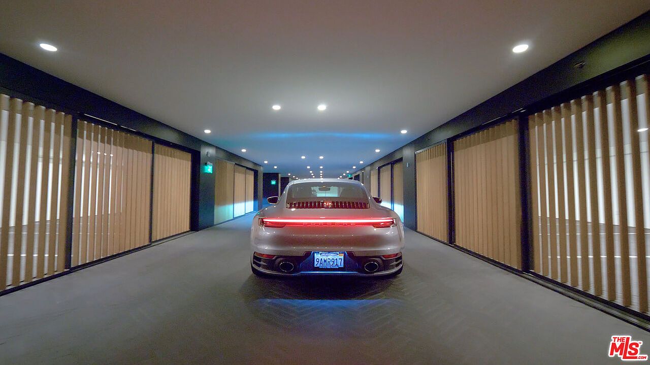 The image showcases a modern hallway with a sleek, gray Porsche parked in the center. The hallway is lined with vertical slat walls, creating a contemporary and minimalist aesthetic. Recessed lighting illuminates the space, highlighting the car and the clean lines of the architecture.