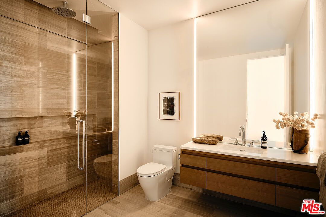 This is a modern primary bathroom featuring a glass-enclosed shower with horizontal wood-look tiles and a rainfall showerhead. The vanity has a light wood finish with a white countertop, a rectangular sink, and a large mirror with integrated vertical lighting. A white toilet sits next to a framed piece of art, and the flooring appears to be light-colored tile, creating a clean and spa-like atmosphere.