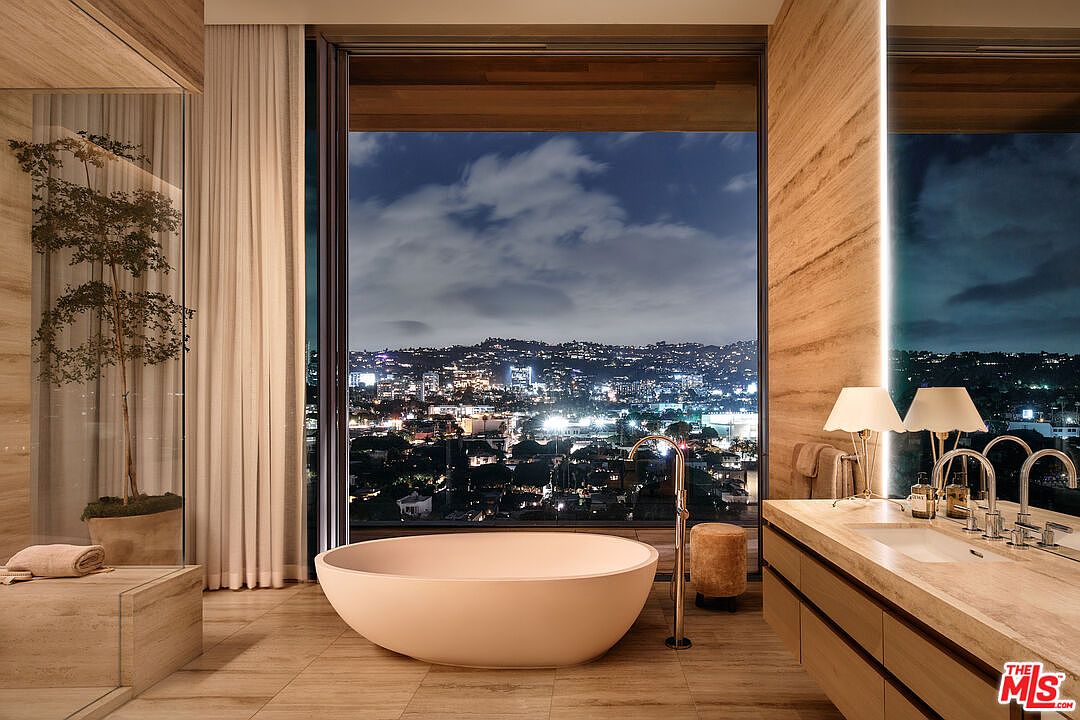 This is a luxurious primary bathroom featuring a freestanding oval bathtub positioned in front of a large window with a stunning city view at night. The bathroom boasts travertine walls and flooring, a modern vanity with dual sinks, and elegant lighting fixtures. A potted tree adds a touch of nature to the space, creating a serene and spa-like atmosphere.