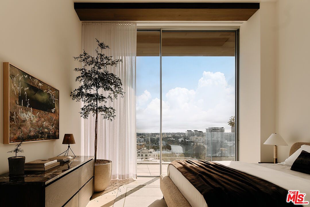This primary bedroom features a large window with city views, complemented by sheer curtains and a decorative tree. A dark wood dresser with artwork and a lamp sits against the wall, while a bed with a brown throw blanket is positioned in the foreground. The room exudes a modern and serene atmosphere.