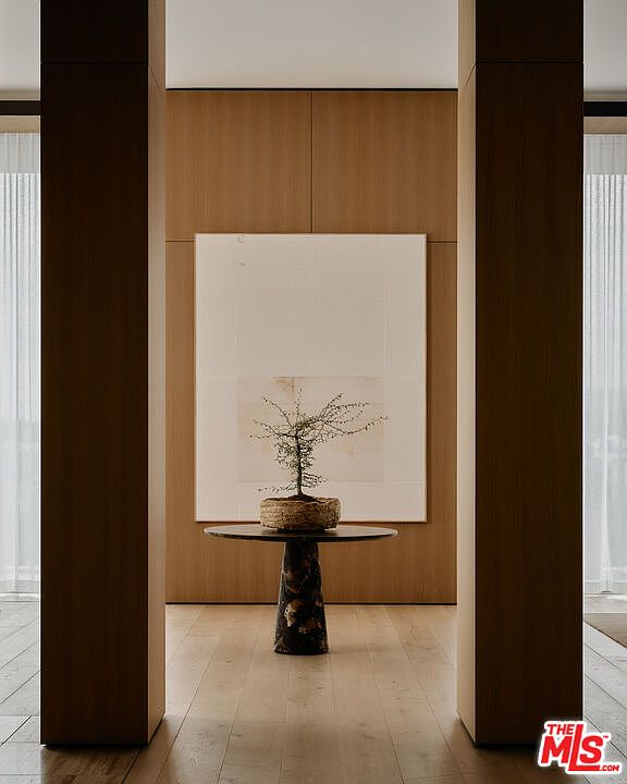This interior shot showcases a modern hallway with a minimalist aesthetic. The hallway is framed by dark wood pillars, leading to a light-filled space with a wooden accent wall and a large piece of artwork above a round table with a small plant. The flooring is light wood, and the overall impression is clean, elegant, and serene.