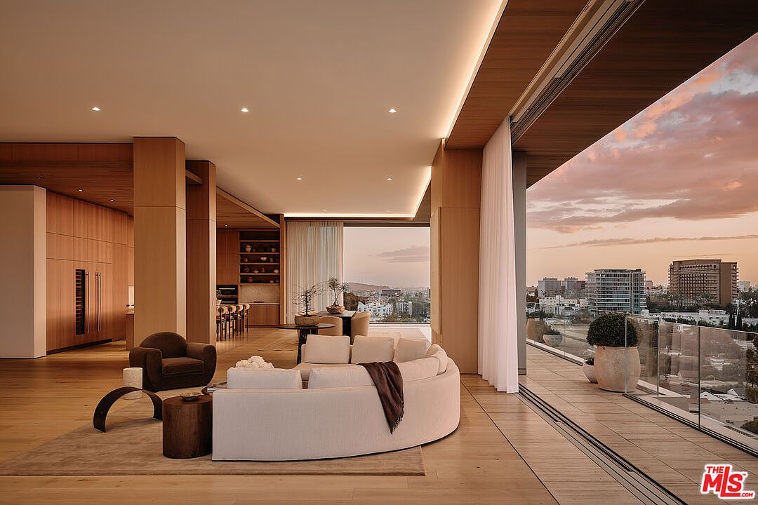 This is a luxurious living room featuring a curved white sofa, a brown armchair, and wooden accents throughout. Large sliding glass doors open to a balcony with city views at sunset, creating a seamless indoor-outdoor living experience. The space is well-lit with recessed lighting and natural light, offering a modern and sophisticated ambiance.