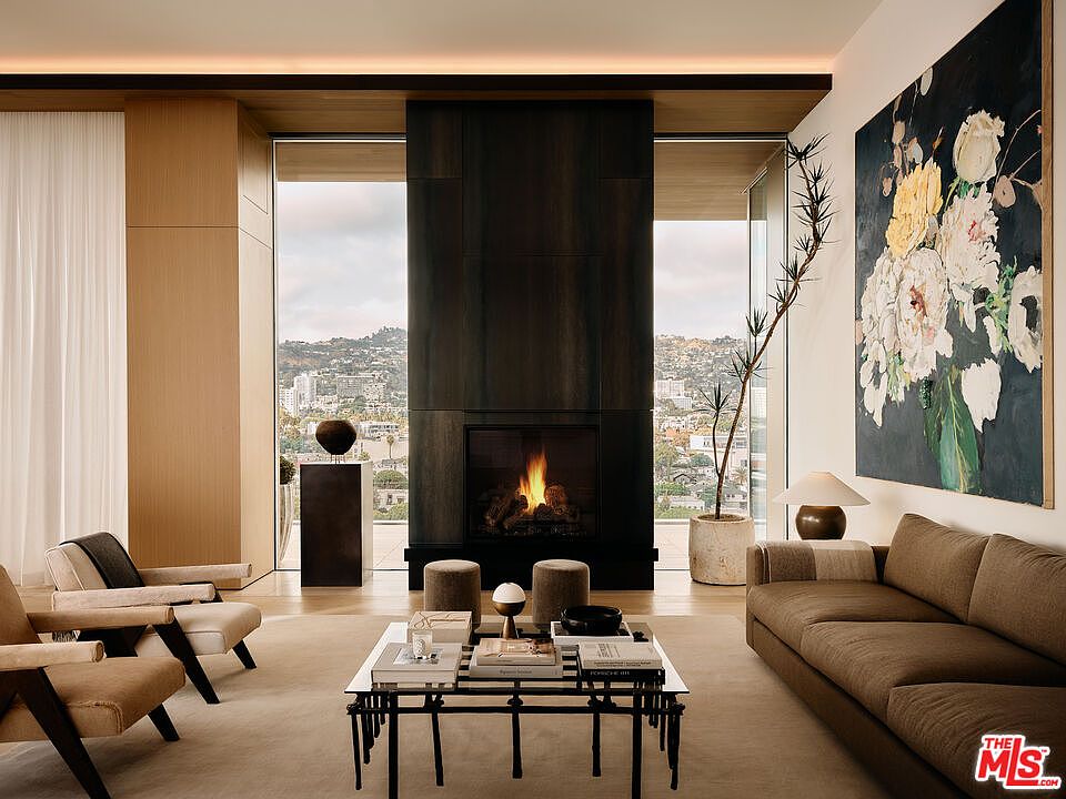 This is a luxurious living room featuring a modern fireplace, a comfortable brown sofa, and two stylish armchairs. A large floral painting adds a touch of elegance, while the floor-to-ceiling windows offer stunning city views. The room is well-lit and spacious, creating a sophisticated and inviting atmosphere.