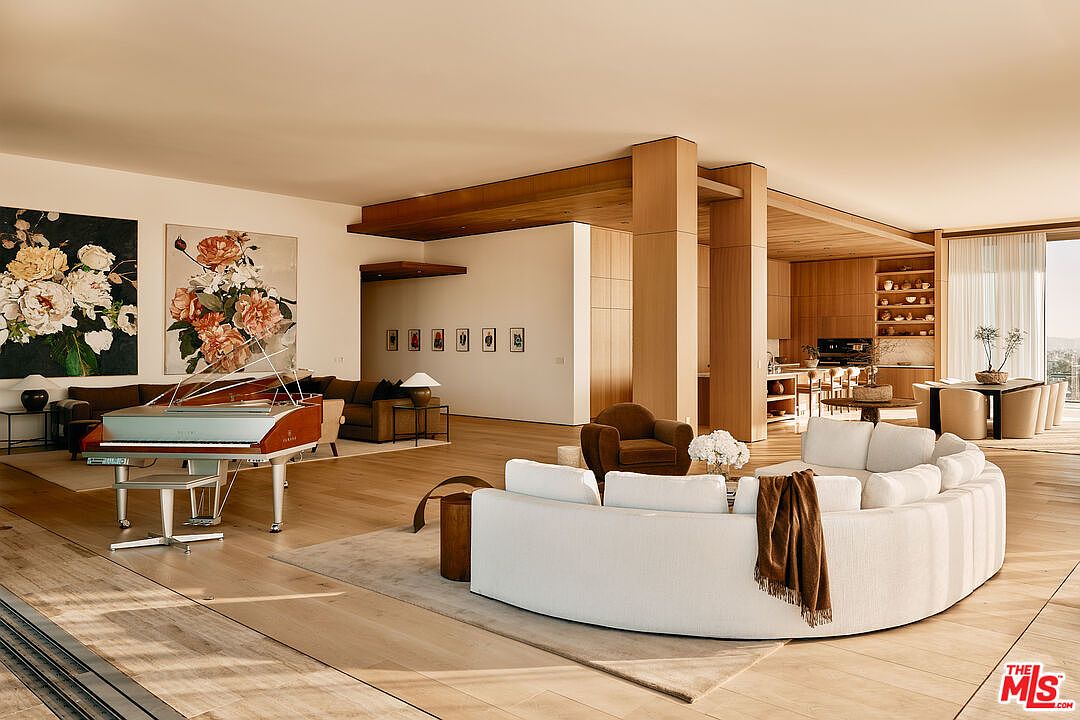 This expansive living room features an open-concept design with light wood flooring and a neutral color palette. A grand piano sits near large floral artwork, while a curved white sofa anchors the seating area. The space flows seamlessly into the kitchen and dining area, creating an inviting and luxurious atmosphere.