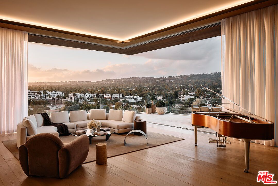This is a luxurious living room featuring a grand piano, a large sectional sofa, and a modern armchair. The room boasts floor-to-ceiling windows that offer panoramic views of the city and hills, seamlessly blending indoor and outdoor living. The hardwood floors and recessed lighting add warmth and sophistication to the space.