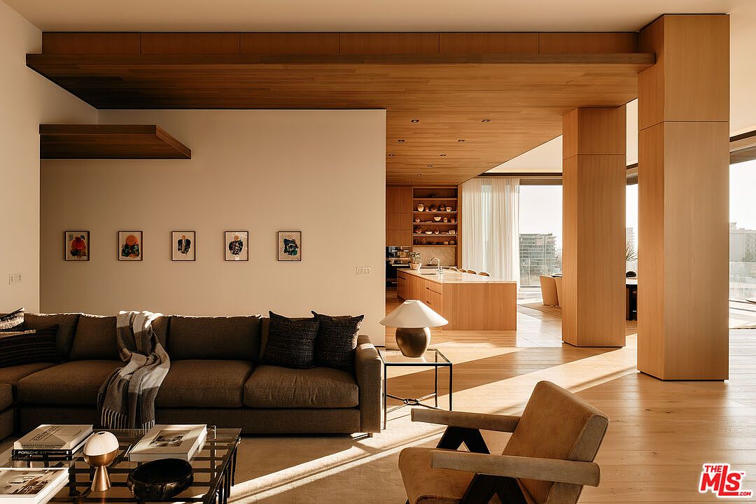 This interior shot showcases a modern living room with a seamless transition to the kitchen area. The living room features a comfortable brown sectional sofa, a glass coffee table, and artwork adorning the wall. The open floor plan, wooden ceiling, and large support beams create a spacious and airy atmosphere, enhanced by natural light streaming through the windows.