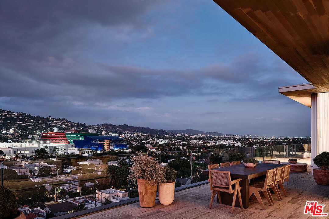 This image showcases a luxurious outdoor patio or deck area with a stunning city view at dusk. A large wooden dining table with chairs is the focal point, complemented by potted plants and a sleek, modern architectural design. The space exudes sophistication and offers an ideal setting for outdoor dining and entertainment.