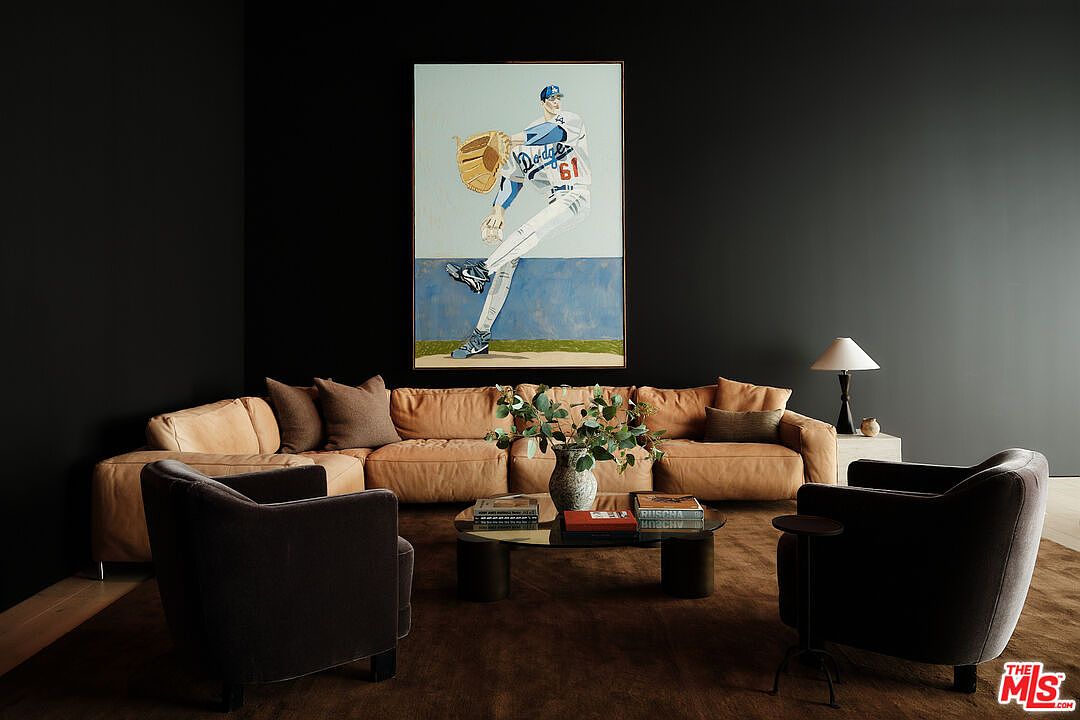 This interior shot showcases a modern living room with dark walls, a large sectional sofa, and two armchairs arranged around a coffee table. A striking painting of a baseball player hangs above the sofa, adding a unique focal point. The room is well-lit with a table lamp, creating a cozy and sophisticated atmosphere.