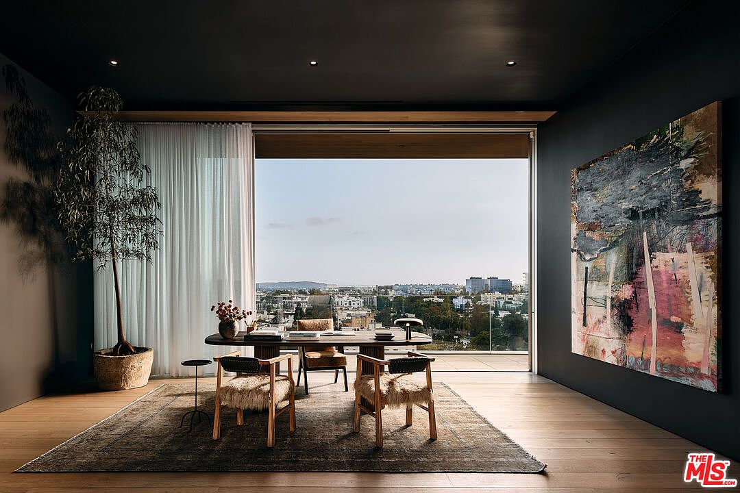 This interior shot showcases a modern office space with a large window offering a panoramic city view. The room features a dark color palette, hardwood floors, and a contemporary rug. A wooden table serves as a desk, surrounded by stylish chairs with fur accents, and a large abstract painting adorns the wall, adding an artistic touch.