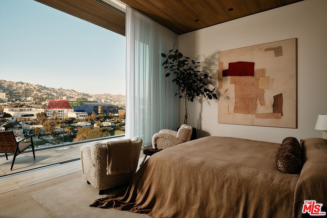 This is a primary bedroom featuring a large bed with a brown cover, two fluffy chairs, and a large window with a view of the city. The room has a modern and minimalist design, with a neutral color palette and an abstract painting on the wall. The overall impression is luxurious and serene.