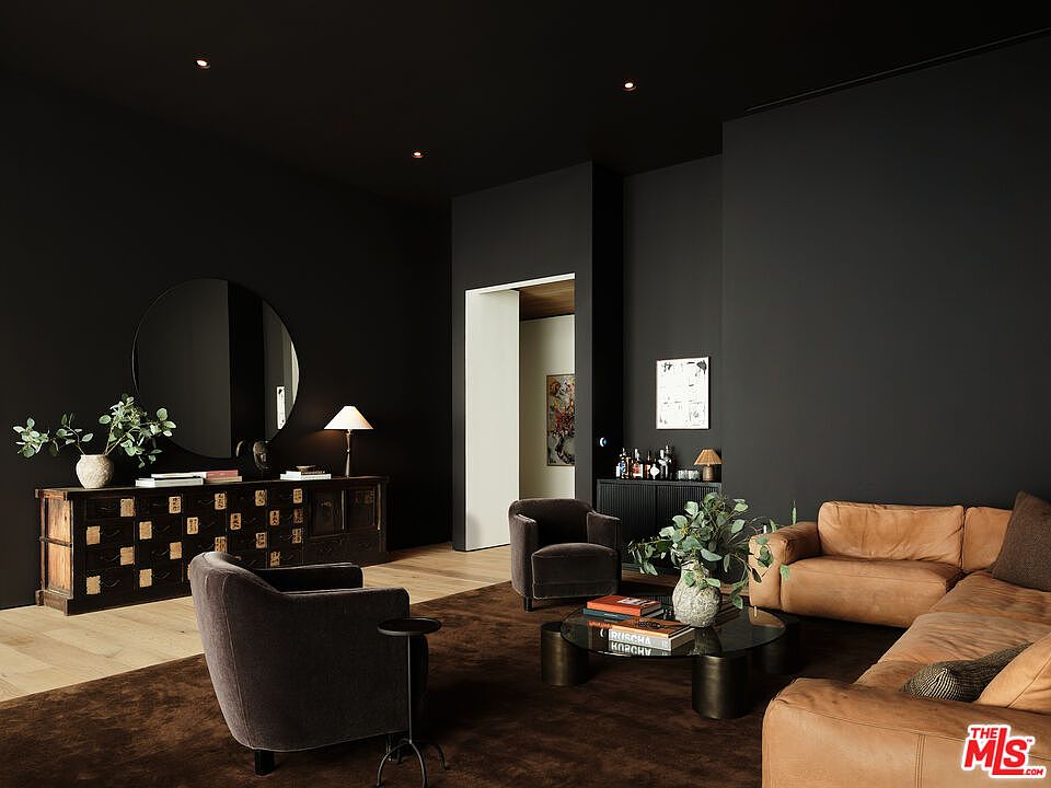 This is an interior shot of a modern living room featuring dark walls and hardwood floors. The room is furnished with a tan leather sectional sofa, two dark armchairs, and a round glass coffee table. A large round mirror hangs above a wooden cabinet, and a doorway leads to another room, creating a sense of depth and flow.