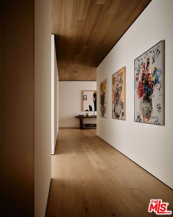 This interior shot showcases a modern hallway with light wood flooring and a wood-paneled ceiling, creating a warm and inviting atmosphere. The white walls are adorned with contemporary artwork, adding a touch of sophistication and visual interest. The hallway leads to a brightly lit area, suggesting a seamless flow between spaces within the home.