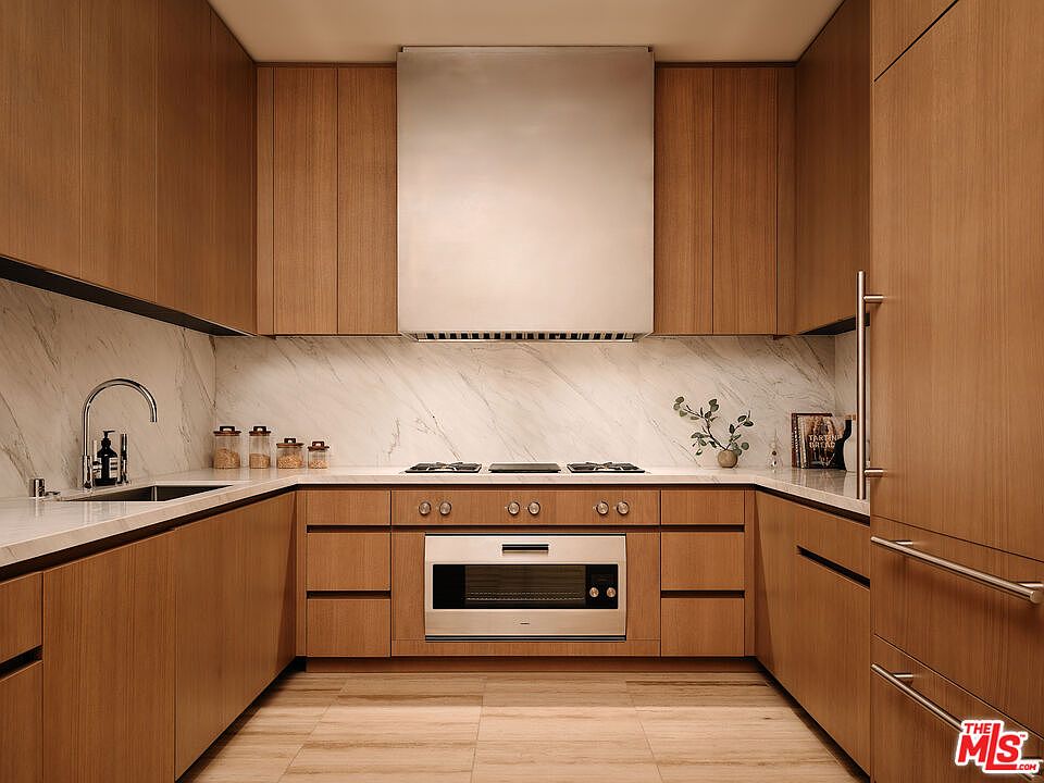 This is a well-lit, modern kitchen featuring sleek, light-wood cabinetry and a white marble backsplash. Stainless steel appliances, including a range hood and built-in oven, add a contemporary touch. The kitchen has a clean and minimalist design, creating a sophisticated and functional space.