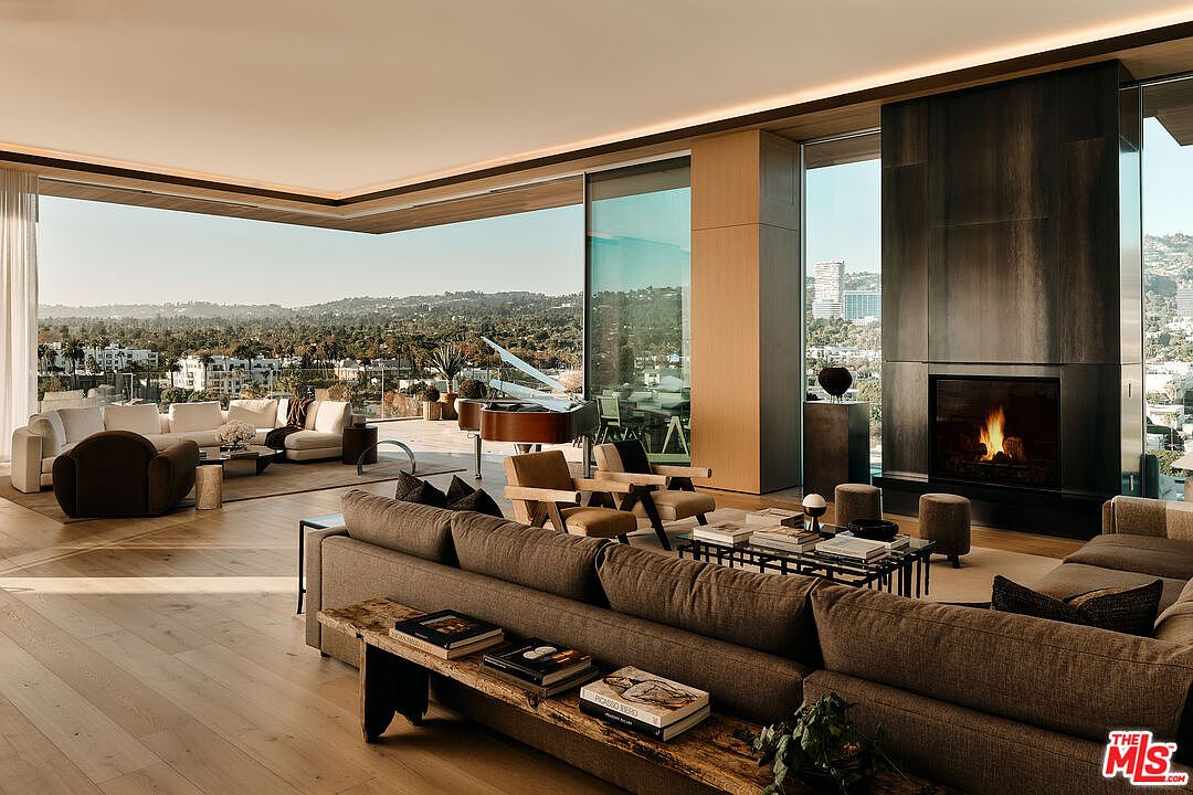 This is a luxurious living room featuring an open floor plan with floor-to-ceiling windows offering panoramic views of the city and surrounding landscape. The room is furnished with a large sectional sofa, modern chairs, a grand piano, and a fireplace, creating a sophisticated and inviting atmosphere. The hardwood floors and neutral color palette enhance the elegance of the space.