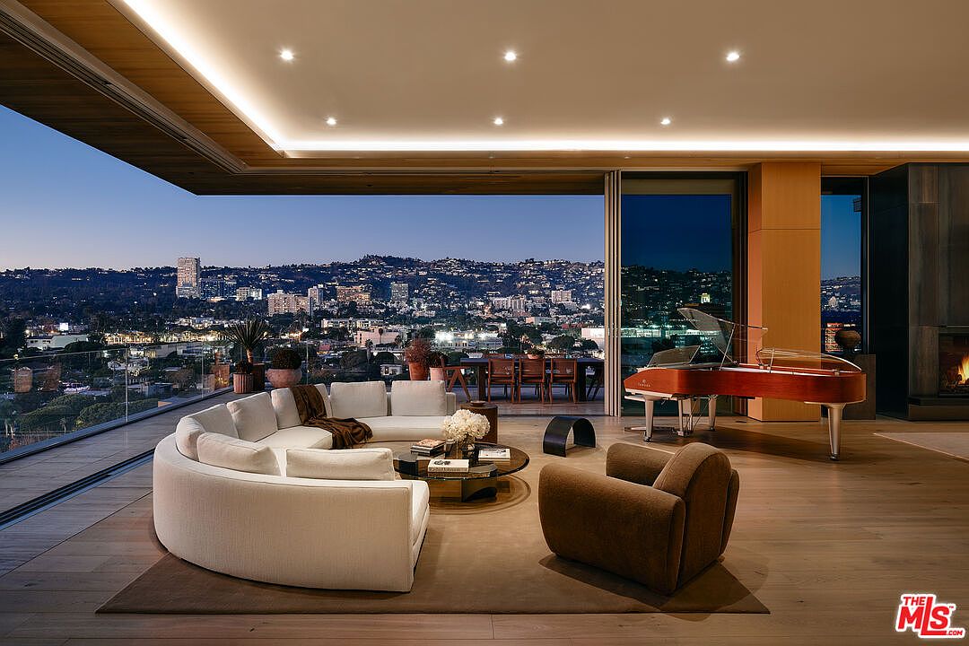 This is a luxurious living room featuring a curved white sofa, a round coffee table, and a brown armchair, all situated on a brown rug. A grand piano sits near the sliding glass doors that open to a balcony with city views at dusk. The room has hardwood floors and recessed lighting, creating a sophisticated and inviting atmosphere.