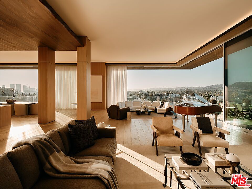 This is a luxurious living room featuring a grand piano, modern furniture, and floor-to-ceiling windows offering panoramic views. The open floor plan and natural light create a spacious and inviting atmosphere. The room is decorated in neutral tones with wood accents, giving it a sophisticated and elegant feel.