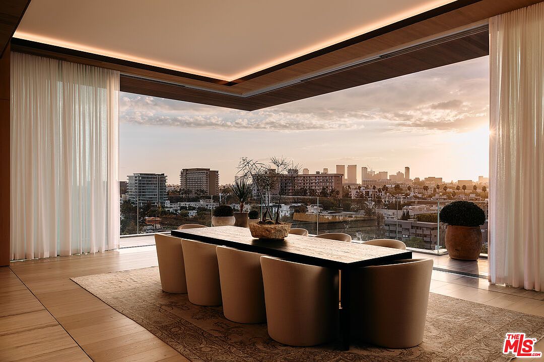 This interior shot showcases a modern dining room with a large, dark wood table surrounded by beige upholstered chairs. Floor-to-ceiling windows offer a panoramic city view at sunset, complemented by sheer white curtains. The room is bathed in warm light, creating an inviting and luxurious atmosphere.