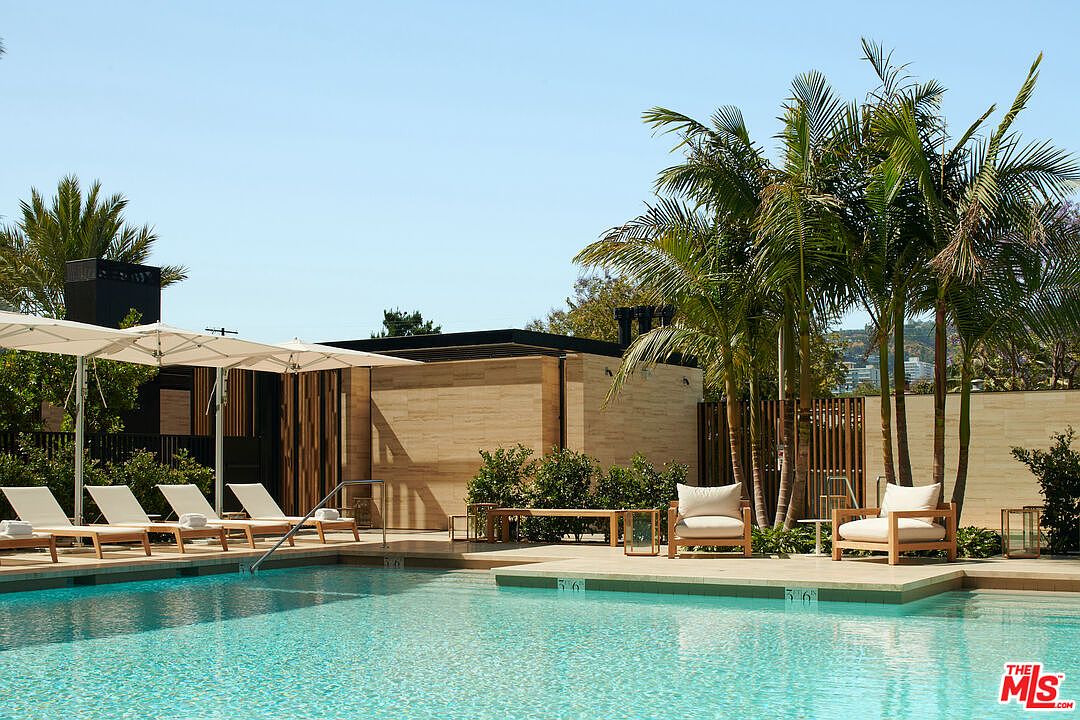 This image showcases a luxurious outdoor pool area, featuring clear turquoise water, modern lounge chairs with umbrellas, and elegant poolside seating. Palm trees add a tropical touch, while the contemporary architecture of the adjacent building enhances the upscale ambiance. The scene evokes a sense of relaxation and high-end living.