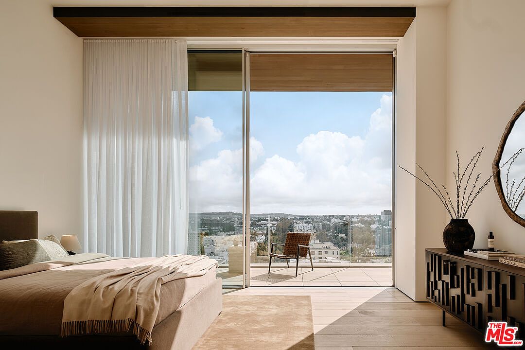 This is a bright and airy primary bedroom featuring a large sliding glass door that opens to a balcony with city views. The room is decorated in neutral tones with a minimalist aesthetic, including a platform bed, sheer curtains, and a modern console table with decorative elements. The hardwood floors and natural light create a serene and inviting atmosphere.
