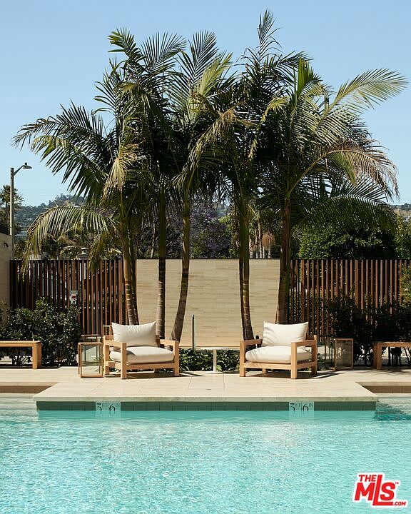 This image showcases a luxurious pool area, featuring clear blue water and a tiled deck with lounge chairs. Palm trees provide shade and create a tropical ambiance, while a wooden fence offers privacy. The scene evokes a sense of relaxation and upscale outdoor living.