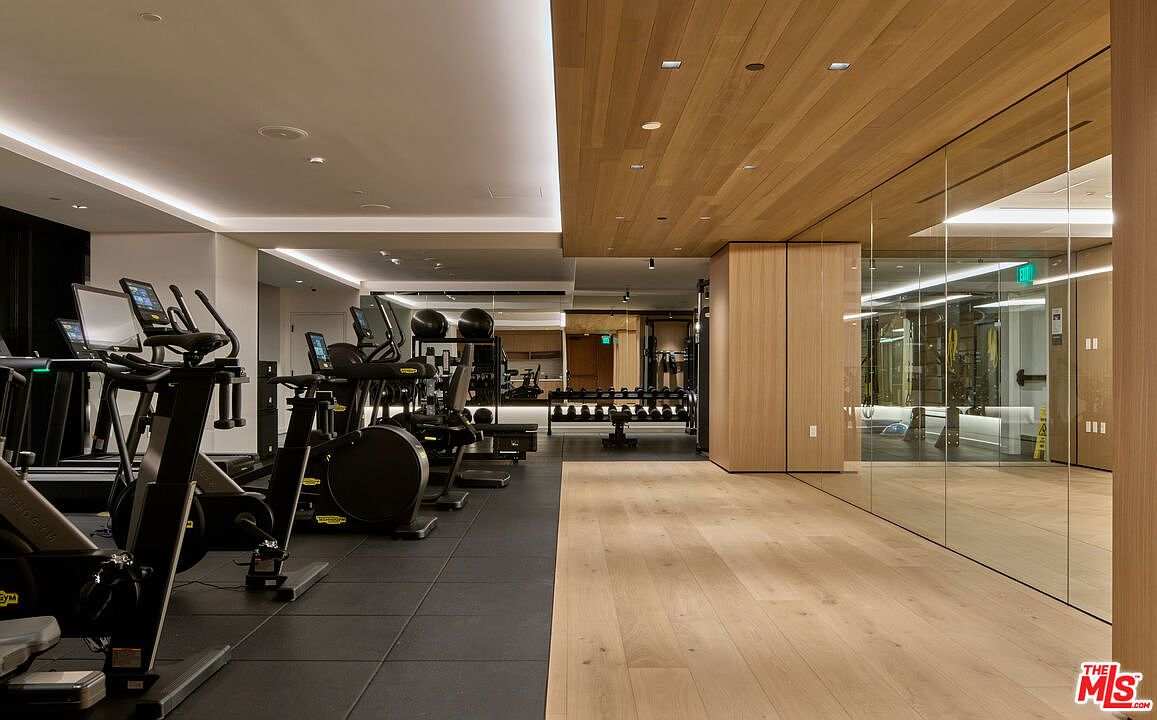This is a modern home gym featuring a variety of exercise equipment including treadmills, stationary bikes, and free weights. The flooring is a combination of dark rubber and light wood, creating distinct zones for different activities. The space is well-lit with recessed lighting and features mirrored walls, enhancing the sense of spaciousness and functionality.