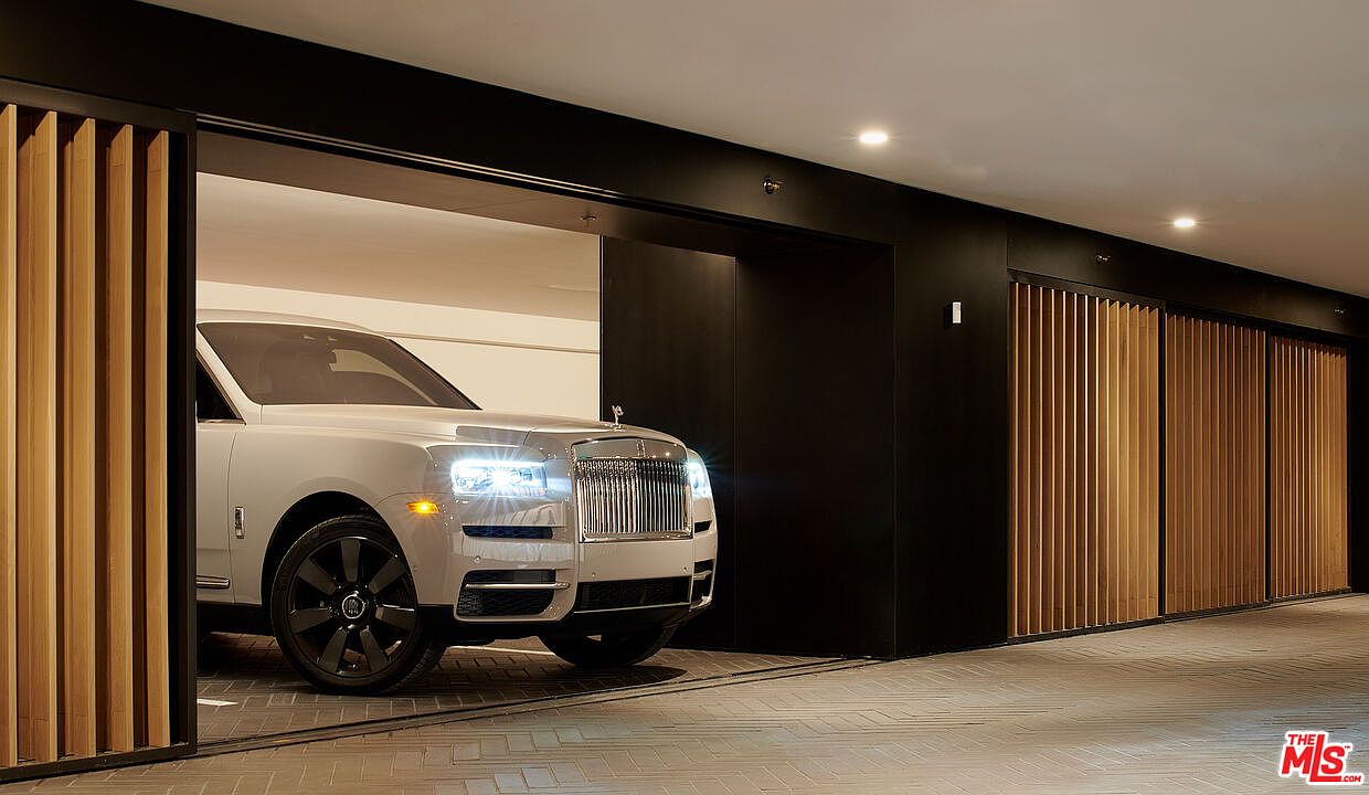 The image showcases a modern garage with a luxury car, likely a Rolls Royce, partially visible inside. The garage features sleek, dark-colored walls and wooden slat doors, creating a sophisticated and contemporary aesthetic. The flooring appears to be brick or tile, adding texture and visual interest to the space.
