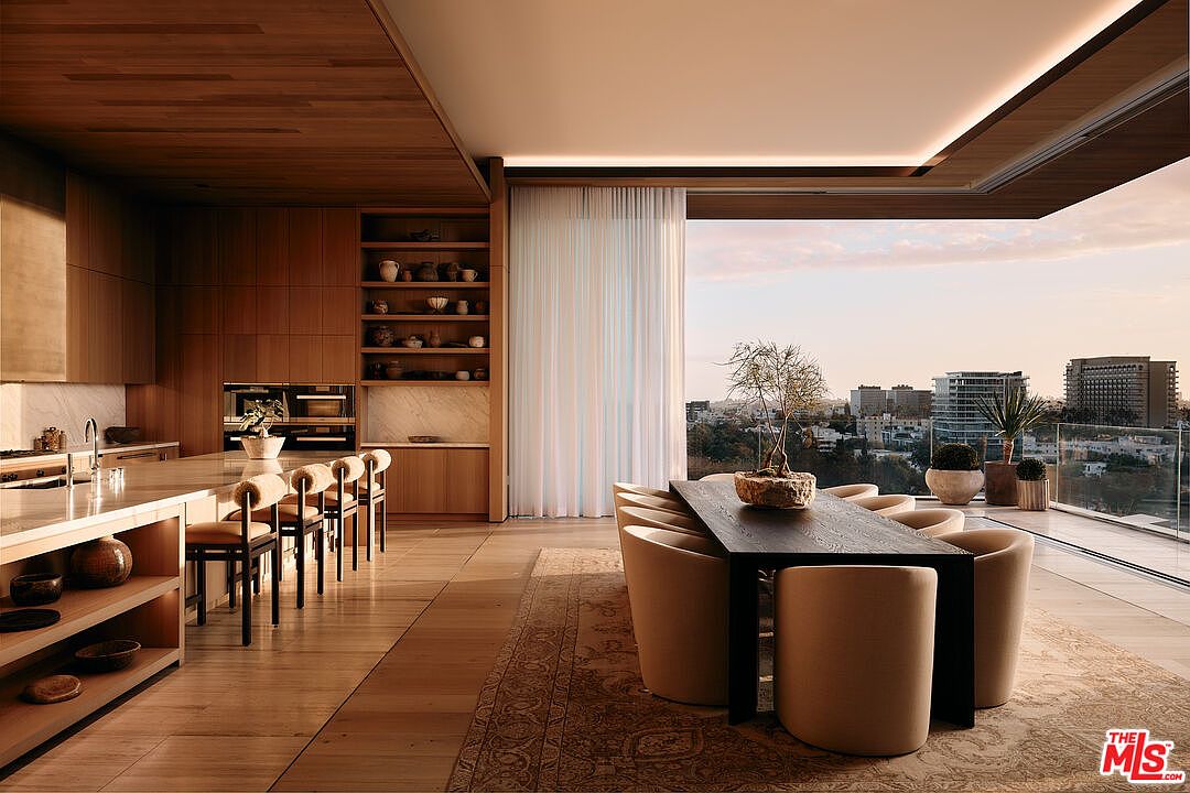 This interior shot showcases a modern dining area with a dark wood table surrounded by beige upholstered chairs, set upon a patterned rug. The space features a sleek, minimalist design with wood flooring, built-in wooden cabinetry, and a large window offering a city view, creating an elegant and inviting atmosphere. A bonsai tree sits on the table, adding a touch of nature.