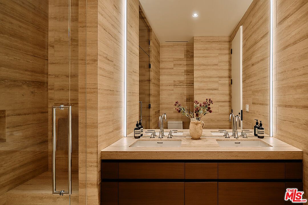 This is a primary bathroom featuring a double vanity with a light-colored countertop and dark wood cabinetry. The walls and shower are tiled in a horizontal pattern with a natural stone look. Large mirrors with integrated lighting enhance the space, and the overall design is modern and luxurious.