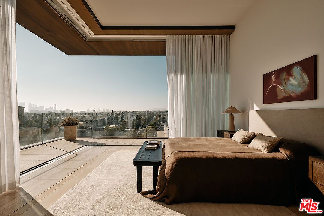 This primary bedroom boasts a stunning city view through a floor-to-ceiling glass wall that opens to a balcony. The room features a neutral color palette, a large bed with a brown cover, and a minimalist aesthetic, creating a serene and luxurious atmosphere. A dark wood bench sits at the foot of the bed, and an abstract painting hangs above the headboard, adding a touch of modern art.