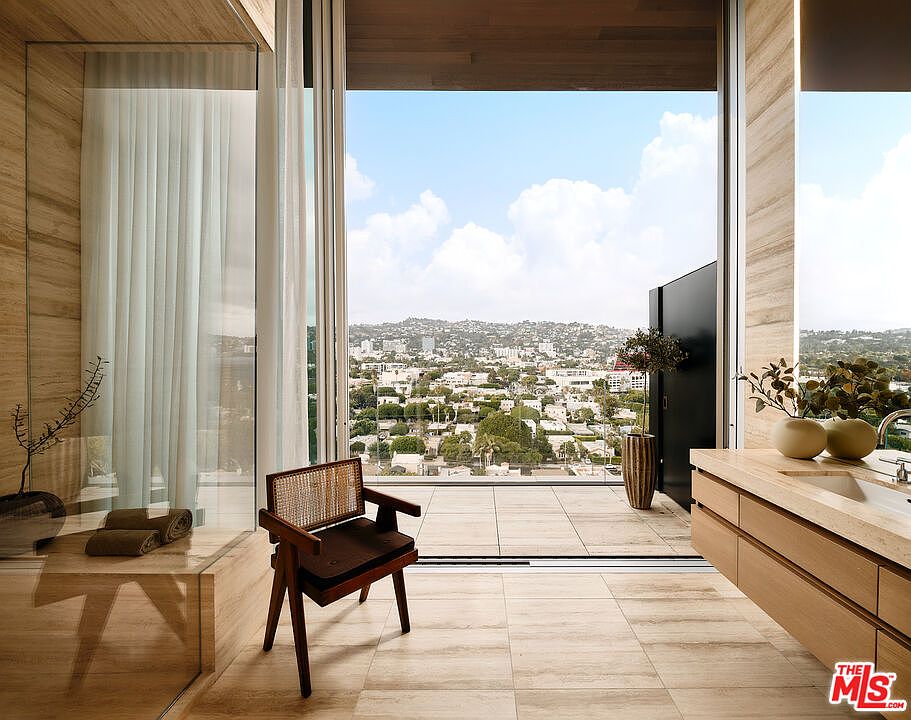 This is a luxurious primary bathroom featuring travertine walls and floors, a glass-enclosed shower, and a modern vanity with dual sinks. A large sliding glass door opens to a balcony with city views, creating an indoor-outdoor living experience. A stylish chair adds a touch of elegance to the space.