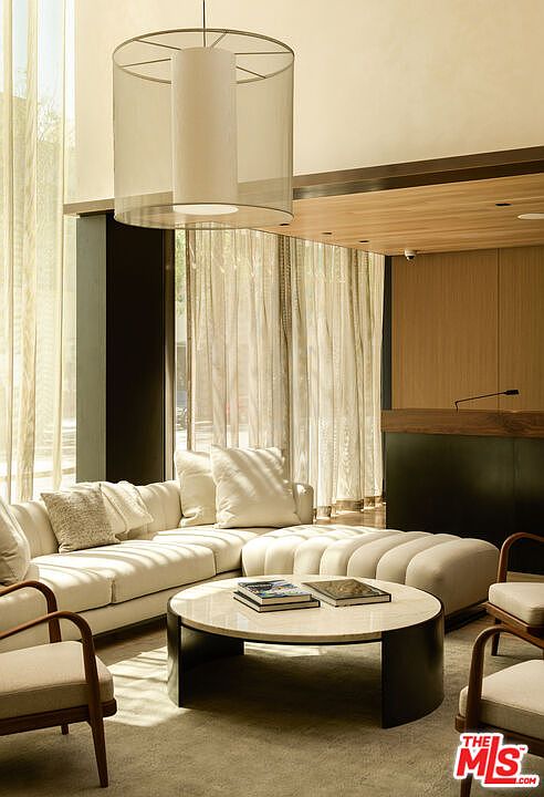 This is an interior shot of a living room featuring a large, off-white sectional sofa with decorative pillows, a round marble coffee table with books, and two armchairs. A modern, cylindrical pendant light hangs from the ceiling, and sheer curtains filter natural light, creating a bright and airy atmosphere. The perspective is from a low angle, emphasizing the room's depth and inviting ambiance.