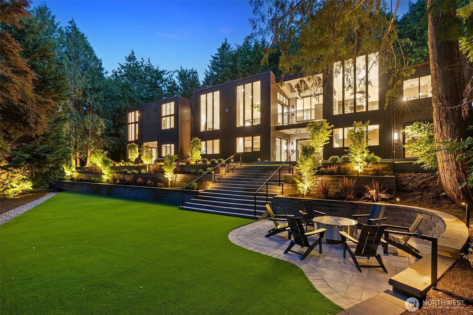 This stunning modern residence features a dark, vertical-paneled exterior with expansive floor-to-ceiling windows that glow warmly against the twilight sky. A tiered landscape design leads from a manicured lawn up a set of stone stairs to the main structure, complemented by a stone patio area with a fire pit and Adirondack chairs. The scene is framed by lush, mature trees, creating a sophisticated and serene atmosphere perfect for evening entertaining.