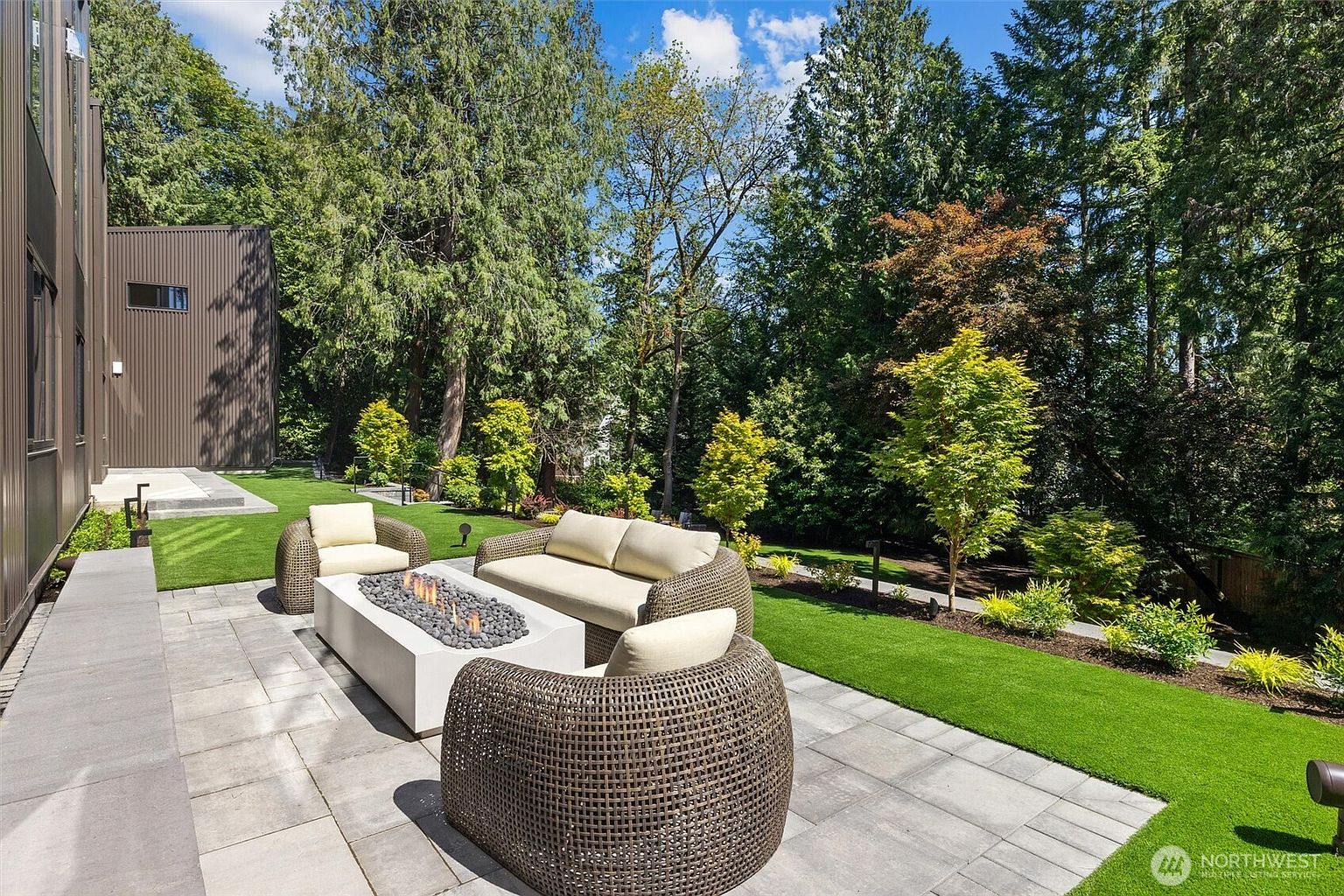 This modern outdoor living space features a sleek, rectangular fire pit surrounded by contemporary wicker patio furniture, set upon a clean-lined stone paver patio. The area seamlessly transitions into a manicured lawn and a lush, wooded backyard, creating a serene and private atmosphere. The perspective captures the inviting seating arrangement against the backdrop of a modern home exterior and dense, mature trees.