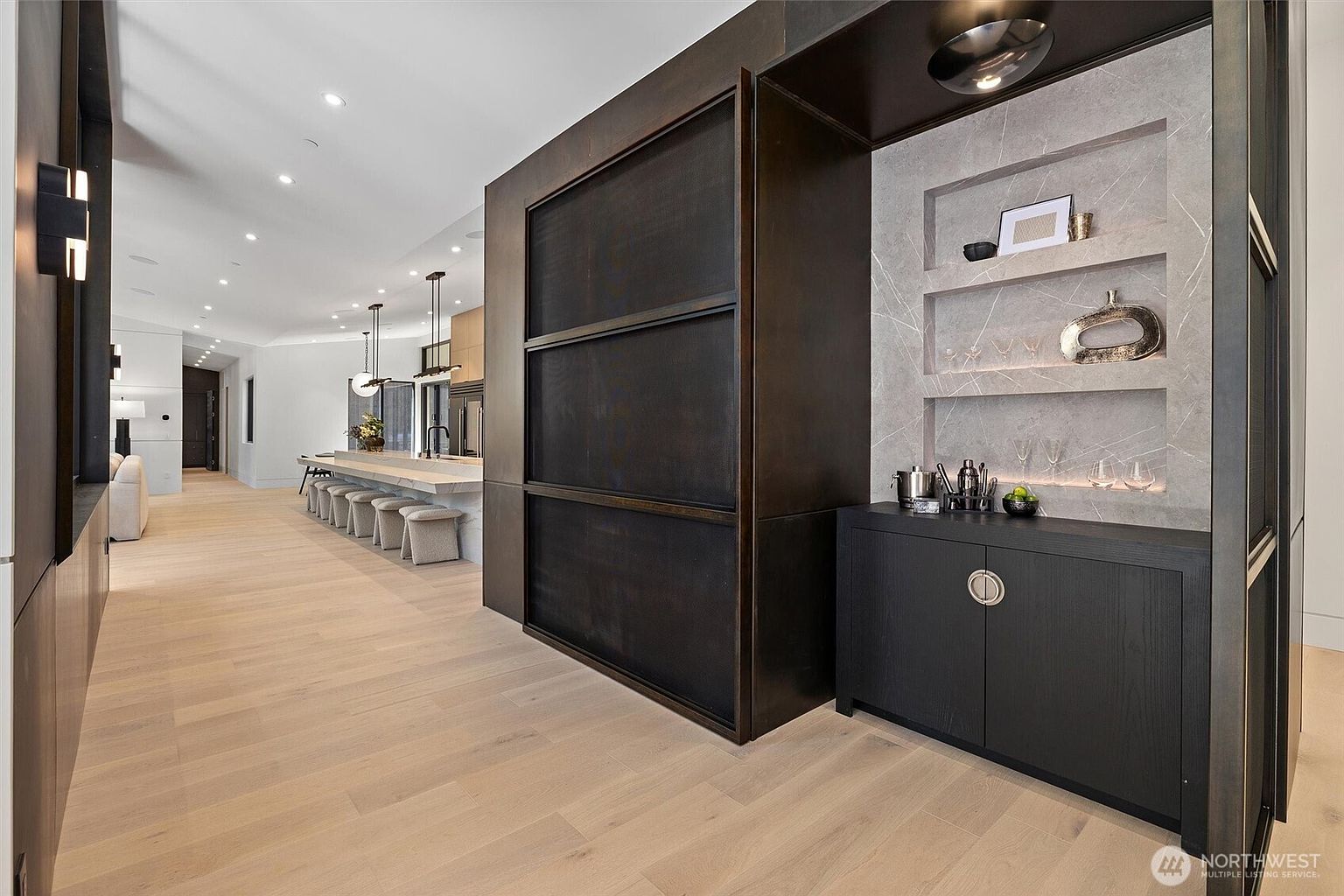 This sophisticated interior view showcases a modern open-concept kitchen and bar area characterized by dark wood paneling and light hardwood floors. A sleek, marble-topped island with bar seating extends into the distance, while a dedicated built-in bar nook with stone shelving and ambient lighting adds a luxurious touch. The perspective emphasizes the clean lines, high-end materials, and seamless flow between the functional kitchen space and the elegant entertaining area.