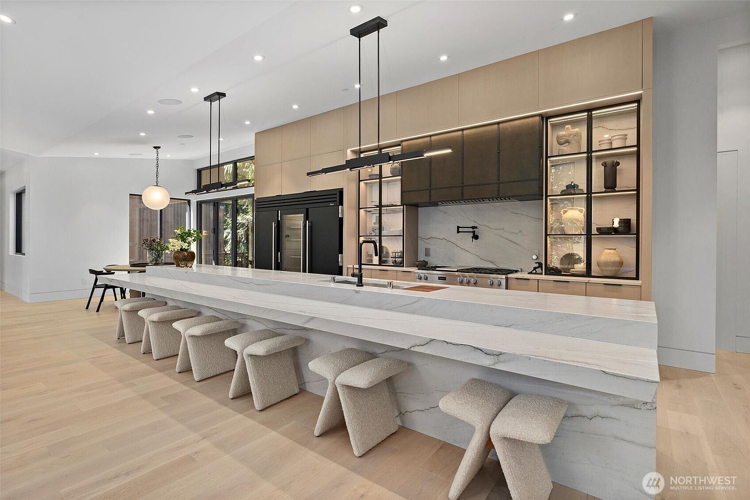 This sophisticated, modern kitchen features a massive waterfall-edge marble island with seating for six, serving as the centerpiece of the open-concept space. The design showcases a seamless blend of light wood cabinetry, dark metal accents, and integrated high-end appliances, complemented by elegant linear pendant lighting. The perspective captures the expansive, airy atmosphere, highlighting the clean lines and luxurious materials that define this contemporary culinary space.