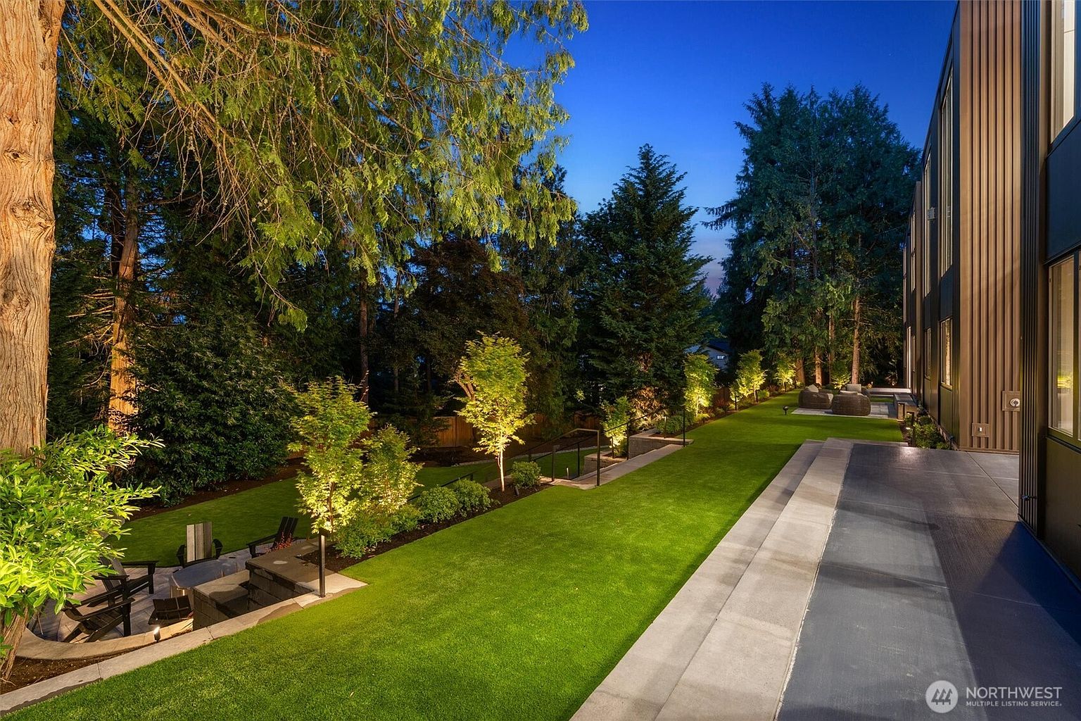 This beautifully landscaped backyard at dusk features a tiered lawn with manicured grass, illuminated by warm landscape lighting that highlights the trees and pathways. A modern, dark-toned home structure runs along the right side, featuring a sleek patio area that transitions into the lush green space. The scene offers a serene and sophisticated atmosphere, perfect for evening relaxation or outdoor entertaining.