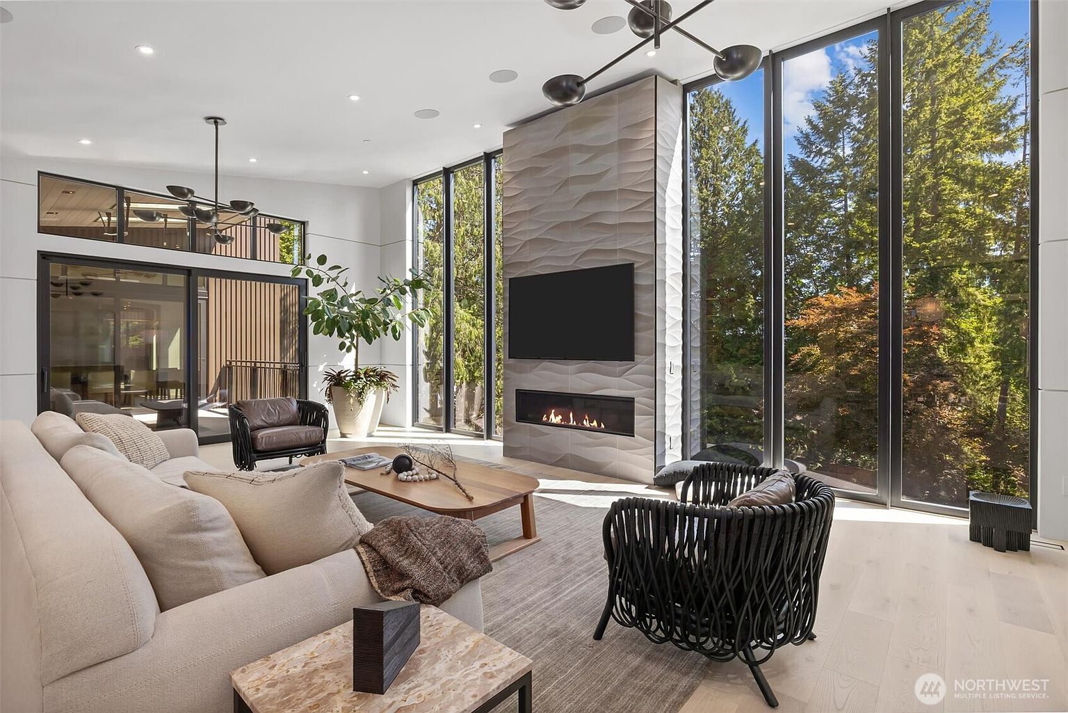 This sophisticated, modern living room features soaring ceilings and floor-to-ceiling windows that frame a lush, wooded outdoor view. A striking, textured stone fireplace serves as the focal point, complemented by a minimalist, contemporary furniture arrangement including a plush sofa and woven accent chairs. The space exudes an airy, luxurious atmosphere, blending high-end architectural design with natural elements.