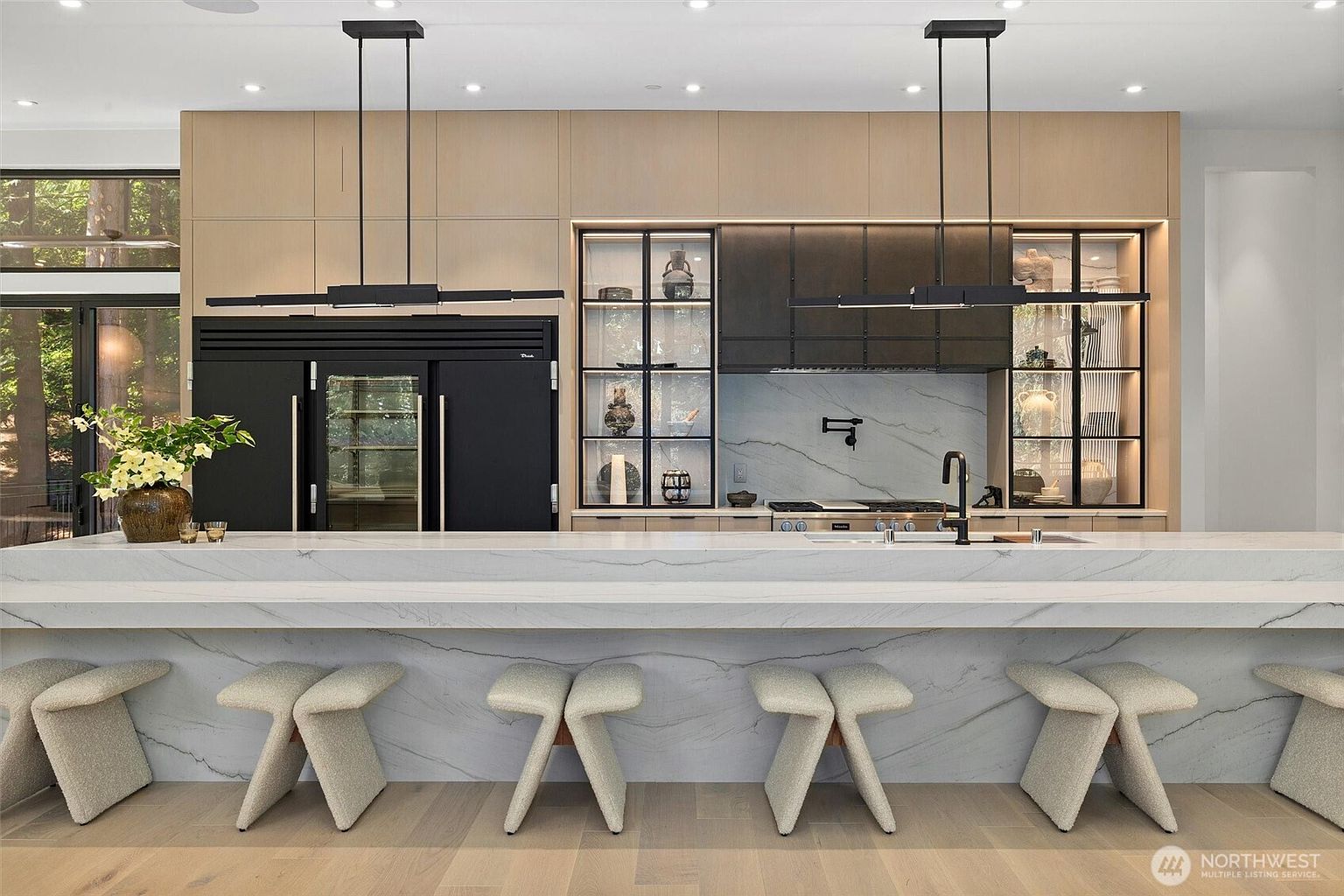 This sophisticated, modern kitchen features a massive waterfall-edge marble island with five textured stools, serving as the centerpiece of the room. The space is defined by sleek, light wood cabinetry, a striking black refrigerator, and illuminated glass-front display cabinets flanking a professional-grade range. Two minimalist linear pendant lights hang above the island, creating a clean, high-end aesthetic that blends luxury with functional design.