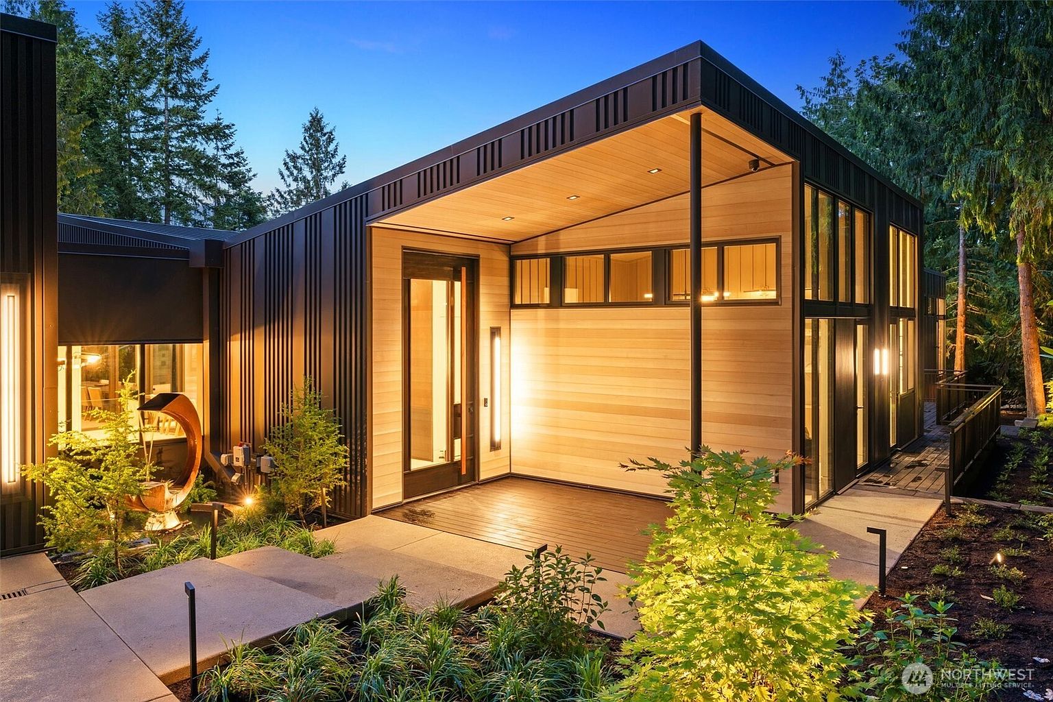 This striking modern entryway features a dramatic blend of dark vertical metal siding and warm natural wood paneling, creating a sophisticated architectural contrast. The covered porch is illuminated by warm, recessed lighting and sleek vertical wall sconces, highlighting the clean lines and contemporary design. A concrete walkway leads to the entrance, surrounded by lush landscaping and a unique sculptural water feature, evoking a serene and high-end atmosphere.