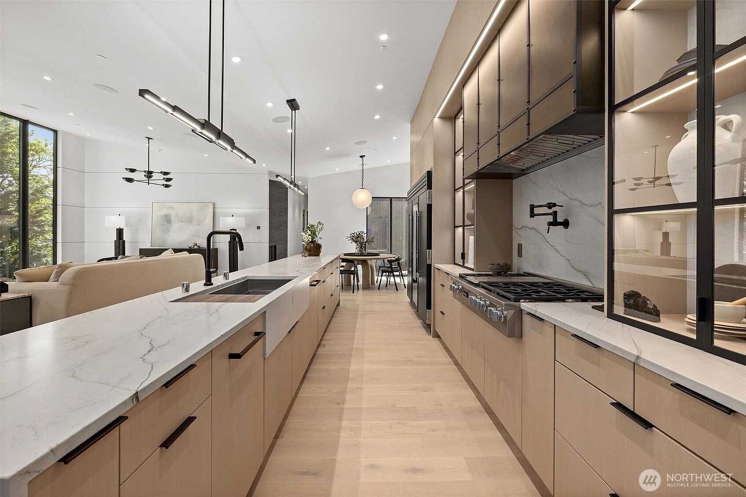 This sleek, modern kitchen features light wood cabinetry, white marble countertops, and high-end stainless steel appliances, including a professional-grade range. The long island serves as a central focal point, complemented by minimalist pendant lighting and an open-concept layout that flows into the living and dining areas. The perspective is a wide-angle shot from the kitchen aisle, emphasizing the clean lines, sophisticated materials, and spacious, airy atmosphere.