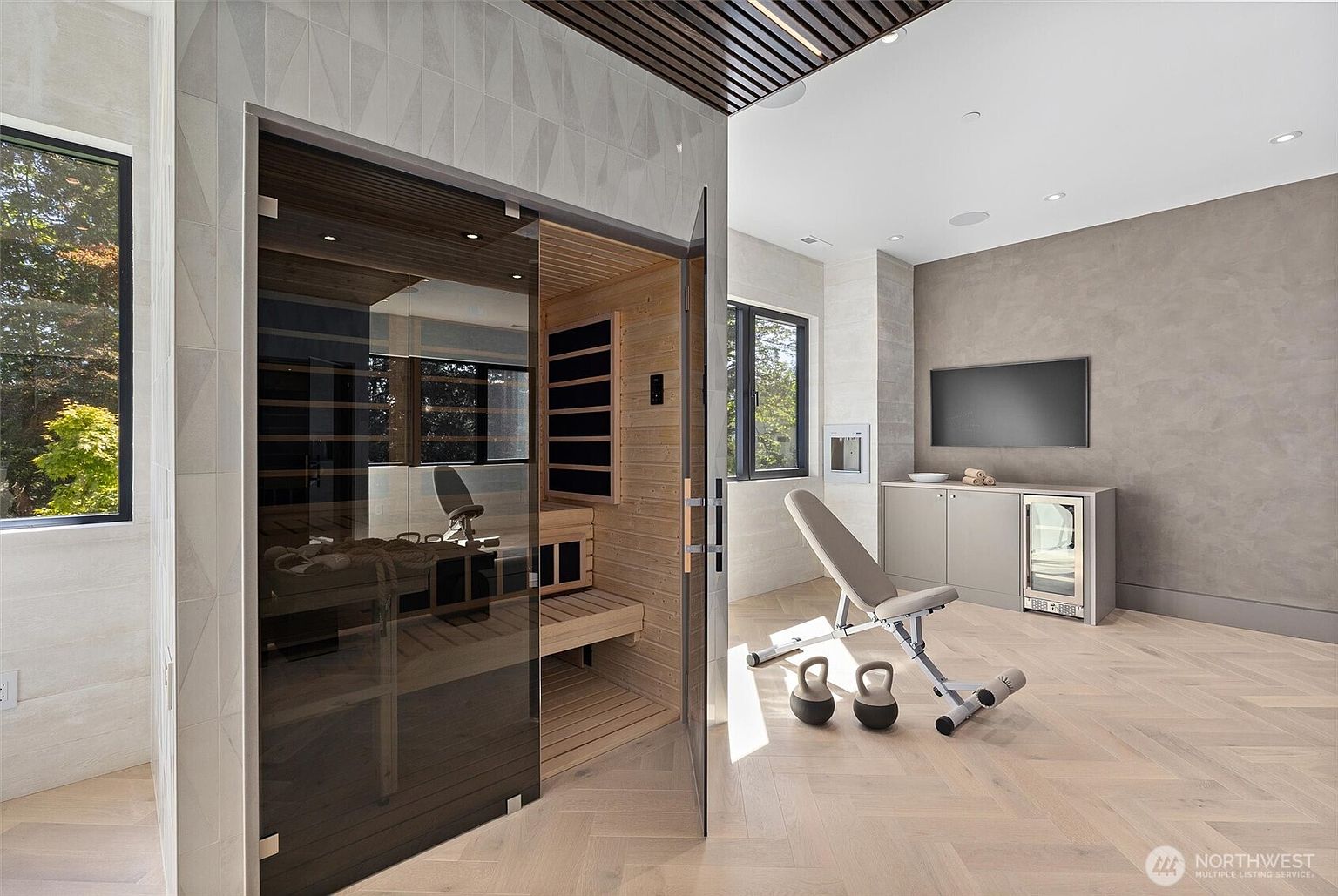 This modern home gym features a sleek, glass-enclosed sauna as its centerpiece, set against a backdrop of light wood herringbone flooring and neutral-toned walls. The space is equipped with a professional-grade adjustable weight bench, two kettlebells, and a built-in refreshment station with a mini-fridge and wall-mounted television. The room exudes a high-end, wellness-focused atmosphere, enhanced by natural light streaming through large windows.