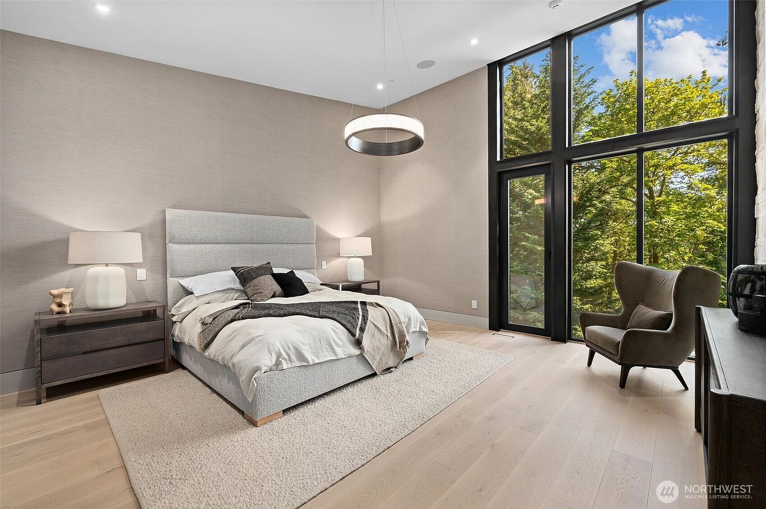 This spacious primary bedroom features a modern, minimalist aesthetic with a large upholstered bed as the centerpiece, complemented by a neutral color palette and textured wallpaper. Floor-to-ceiling windows flood the room with natural light and provide a serene view of the surrounding forest, while a contemporary circular chandelier adds a touch of elegance. The room is furnished with sleek nightstands and a comfortable wingback chair, creating a tranquil and sophisticated retreat.