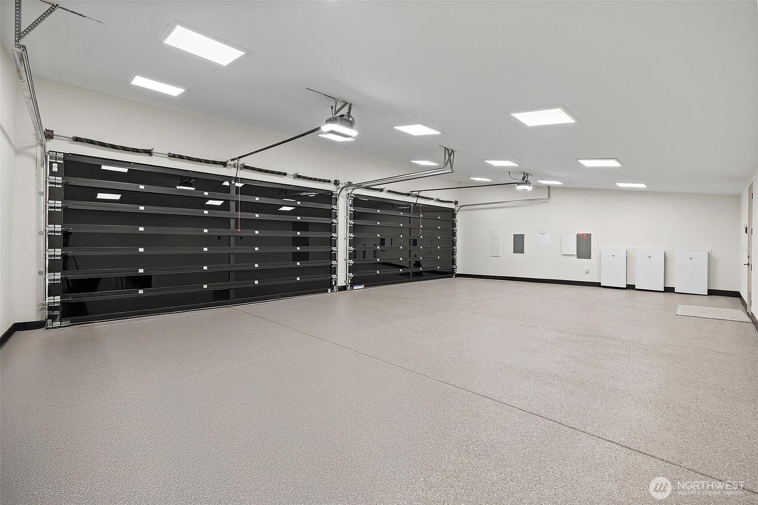 This spacious, modern garage features two large, black-framed glass sectional doors that provide a sleek, industrial aesthetic. The interior is finished with a clean, light-colored epoxy floor and bright recessed ceiling lighting, creating a pristine and professional environment. Along the back wall, several white wall-mounted battery storage units are neatly installed, highlighting the garage's modern utility and energy-efficient design.