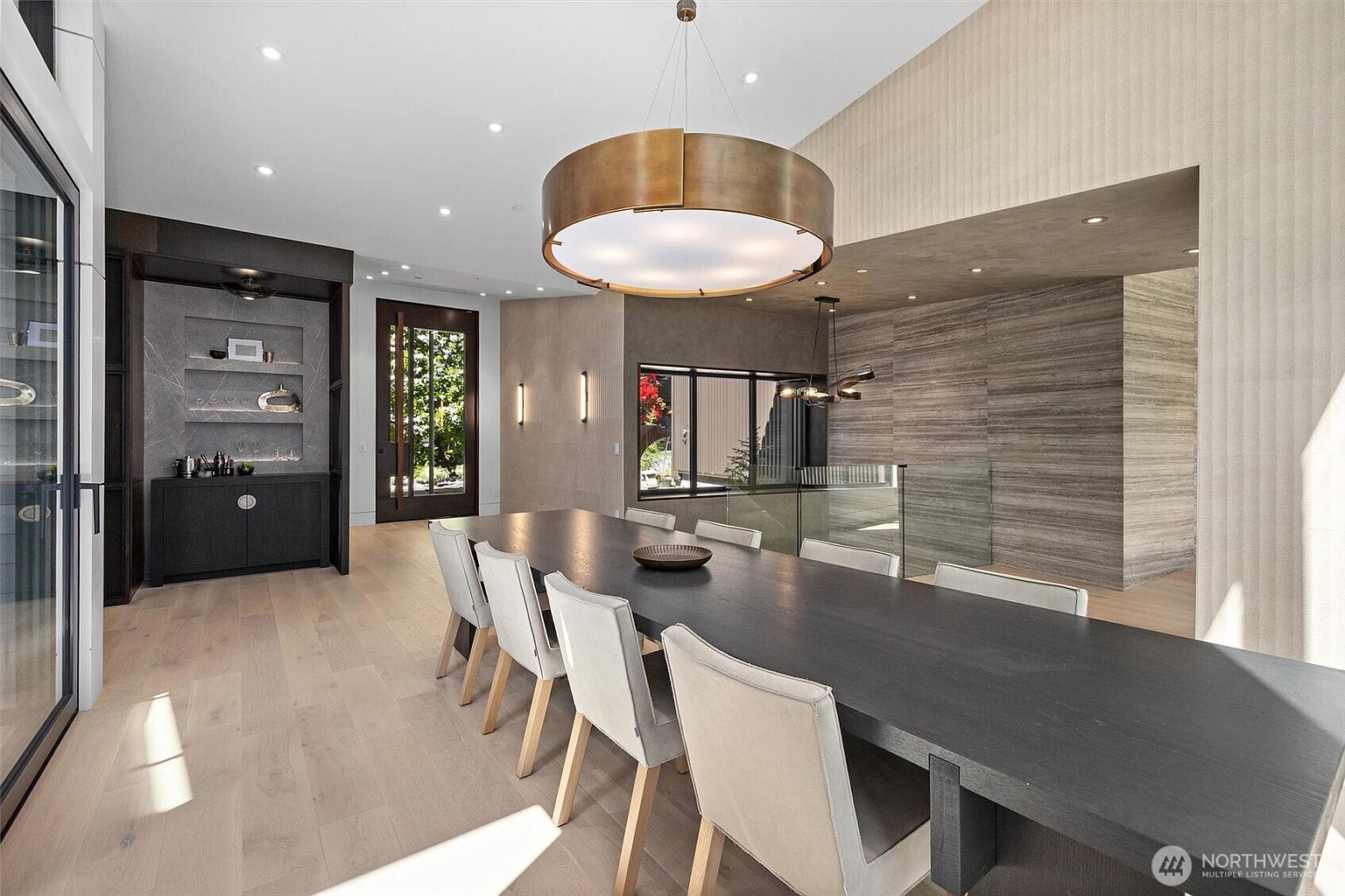 This sophisticated dining room features a long, dark wood table surrounded by modern, light-upholstered chairs, set beneath a striking, oversized brass drum pendant light. The space is defined by high ceilings, light hardwood flooring, and a textured accent wall, with a built-in wet bar area adding a touch of luxury. The perspective captures the open-concept layout, highlighting the seamless transition between the dining area and the adjacent staircase with its glass railing.
