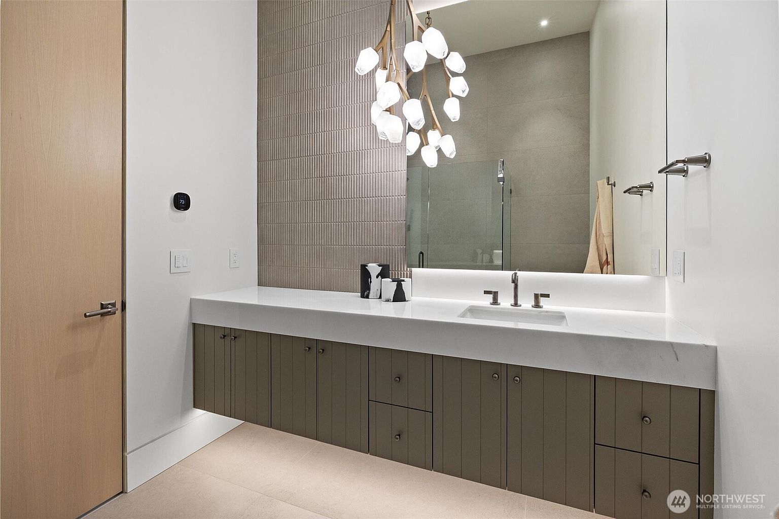 This modern, high-end bathroom features a floating vanity with vertical wood-paneled cabinetry and a sleek white countertop. A large, frameless mirror reflects a unique, branch-like chandelier, while the wall behind the vanity is accented with textured, vertical-ribbed tiles. The space is illuminated by integrated under-cabinet lighting and recessed ceiling lights, creating a sophisticated and serene atmosphere.
