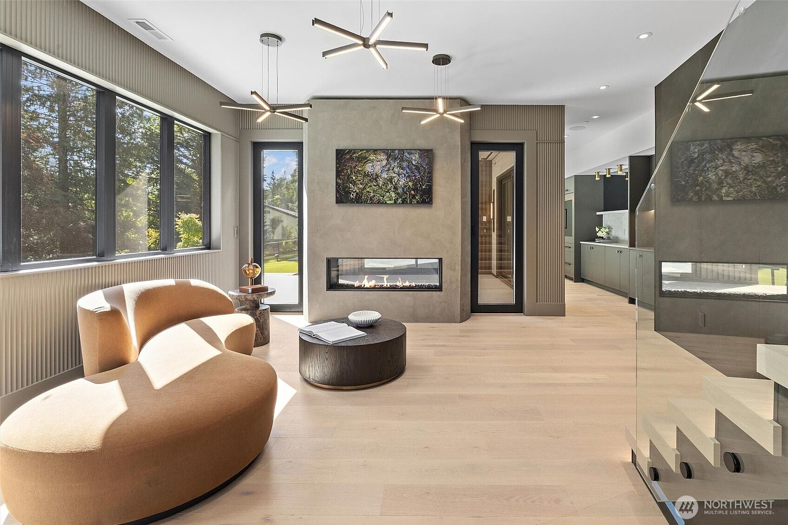 This sophisticated living room features a contemporary, curved tan sofa and a dark wood circular coffee table set against a backdrop of light hardwood floors and textured wall paneling. A sleek, linear fireplace is embedded into a central concrete-style feature wall, topped with abstract artwork and flanked by floor-to-ceiling windows and glass doors. The space is illuminated by modern, geometric pendant lighting, creating an airy and luxurious atmosphere that flows seamlessly into the adjacent kitchen.